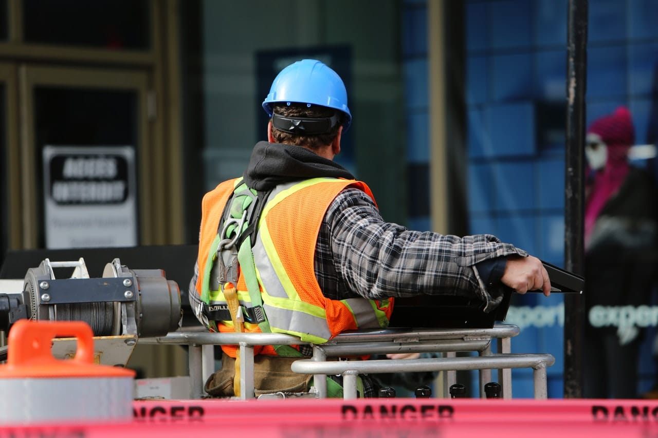 Homme de dos dans une nacelle de chantier, portant un casque de protection bleu, une chemise à carreaux et un gilet orange