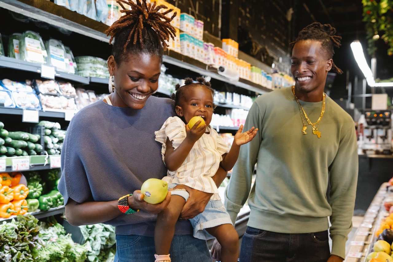 Au rayon fruit d'un supermarché une femme noire souriante porte sa fille à côté d'un homme noir souriant