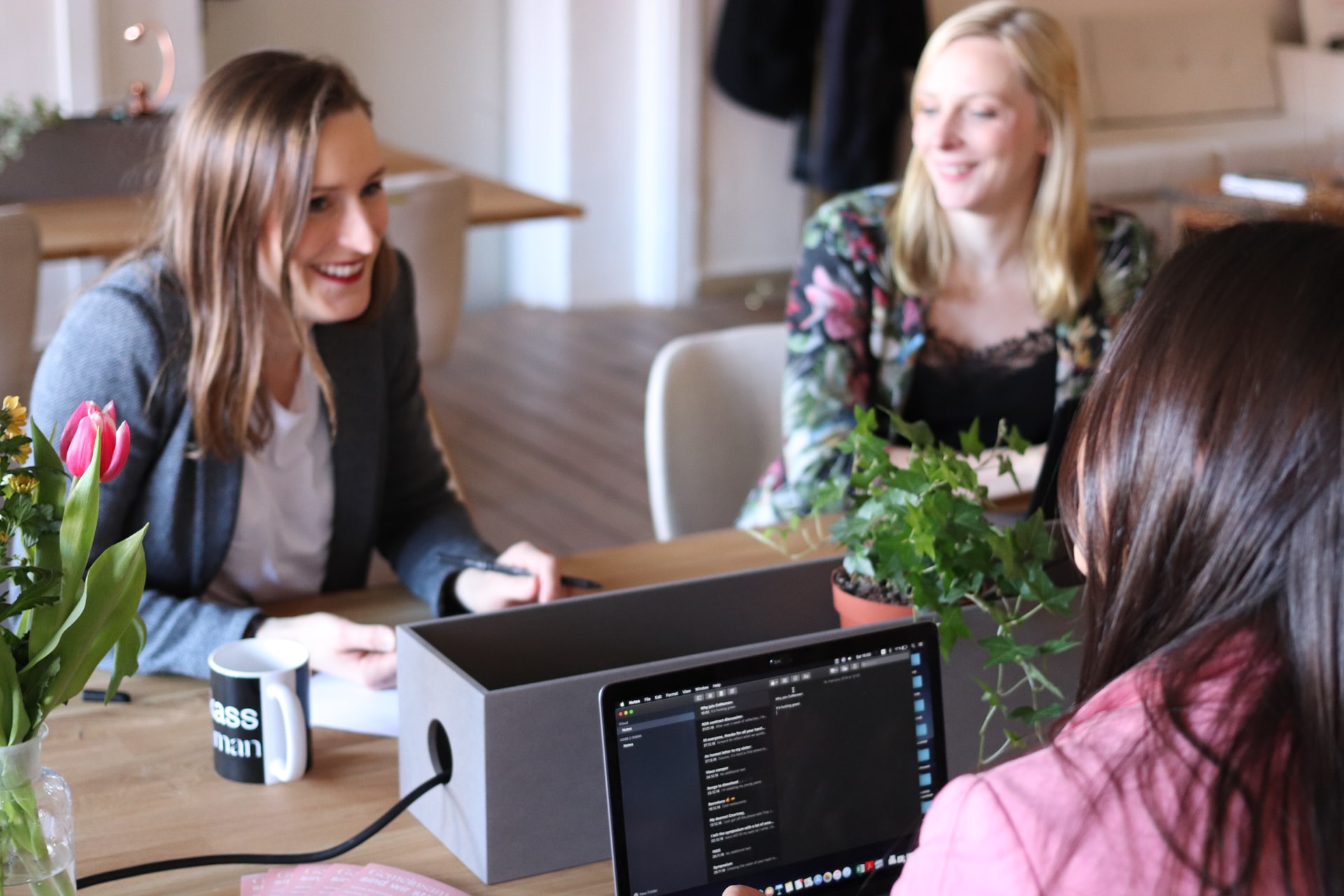 Trois femmes blanches discutant assises autour d'une table devant leurs ordinateurs