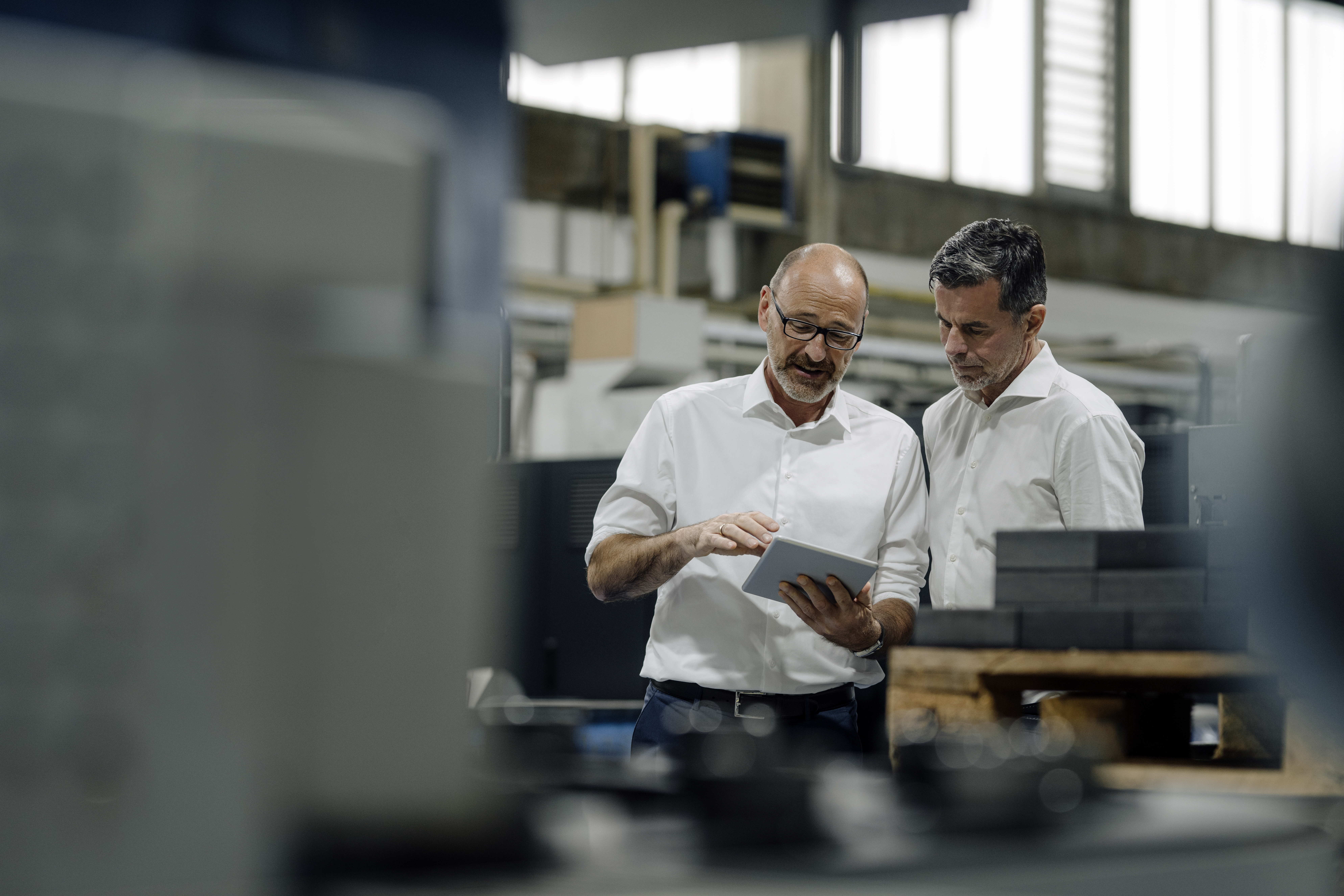 Deux hommes blanc côte à côte dans une usine, celui de gauche montre une tablette à celui de droite