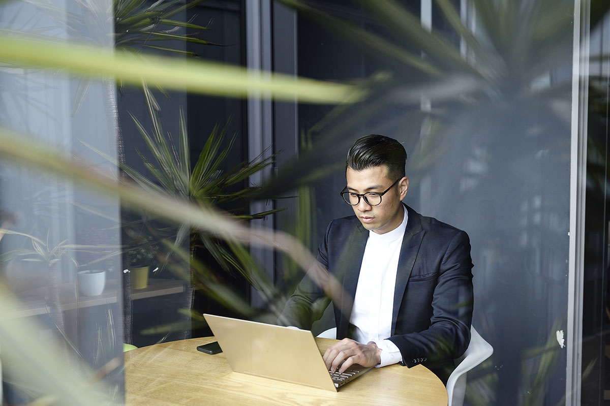 Un homme brun asiatique portant des lunettes, une veste noire et un tshirt blanc assis face à un ordinateur portable