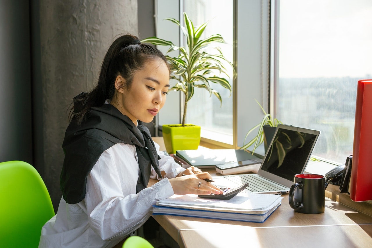 Une femme asiatique assise à un bureau face à une fenêtre tape sur une calculatrice posée à côté d'un ordinateur et d'une tasse