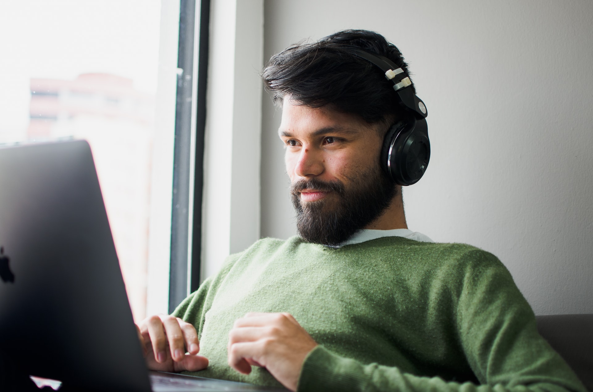 Un homme brun à la peau mate portant un pull vert portant un casque audio assis devant son ordinateur