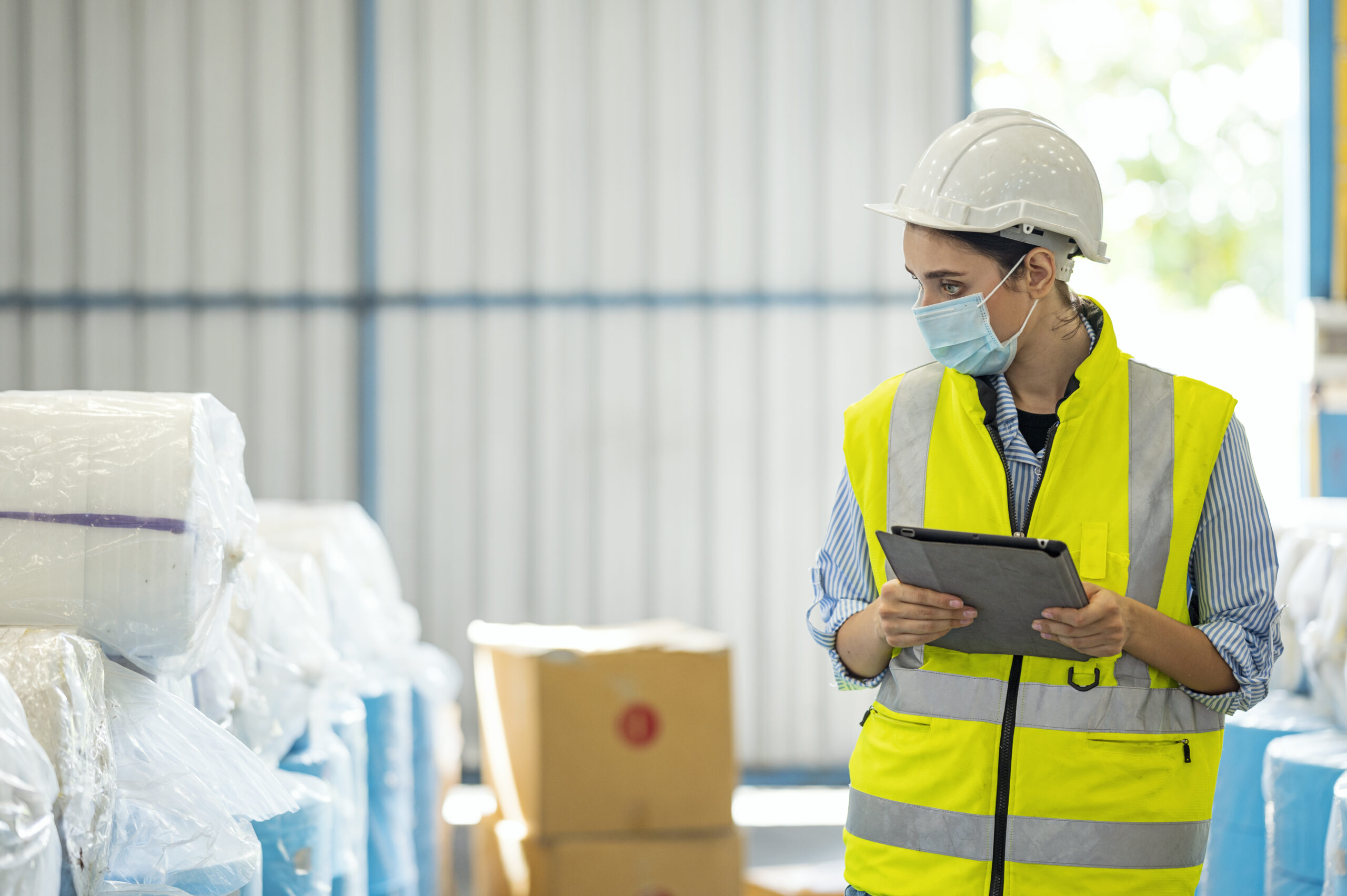 Dans un entrepôt une femme blanche portant un casque et un gilet jaune tient une tablette à la main