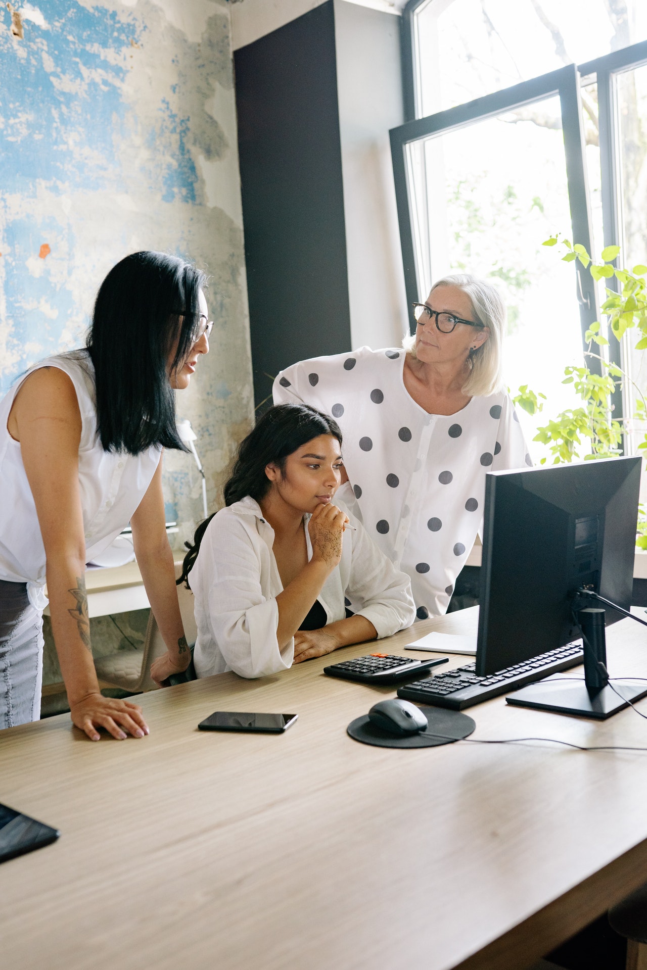 Une femme à la peau mate assise à son bureau entre une femme brune et une femme aux cheveux blancs debout