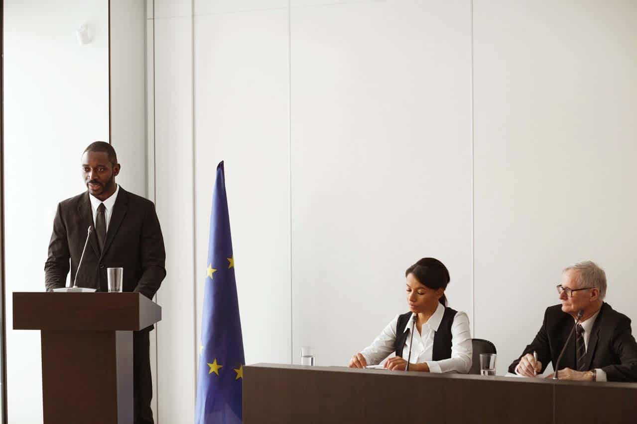 Un homme noir parle à un pupitre devant un drapeau européen à côté d'une femme à la peau mate et d'un homme aux cheveux blancs